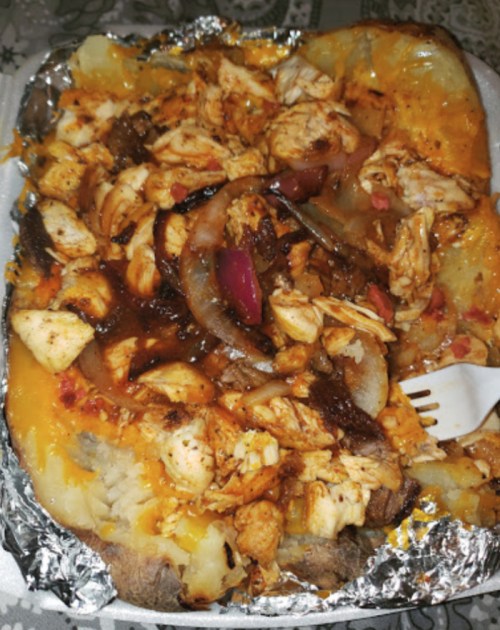 A loaded baked potato topped with shredded chicken, onions, and cheese, served in aluminum foil.