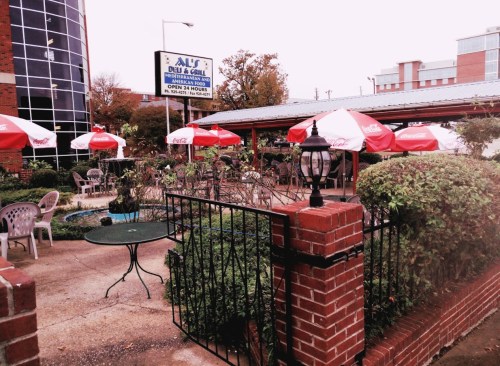 Outdoor seating area of a restaurant with red umbrellas, surrounded by greenery and a small pond.
