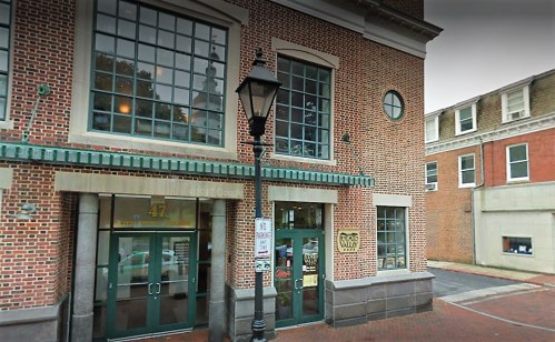 Brick building with large windows, green awning, and a streetlamp outside. Nearby buildings visible in the background.