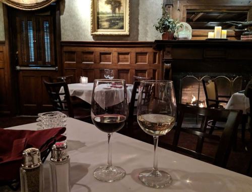 Two wine glasses, one red and one white, sit on a table in a cozy restaurant with a fireplace in the background.