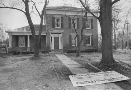 Black and white photo of a historic brick house with a sign in front, surrounded by bare trees and a dirt path.
