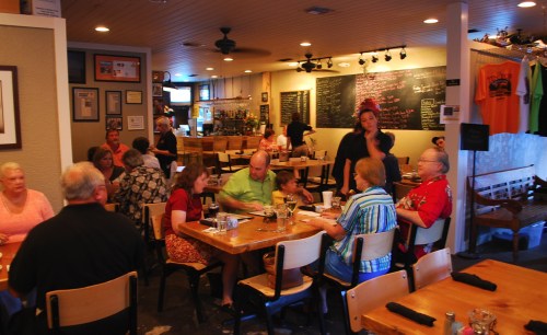 A busy restaurant scene with diners at tables, a server interacting with guests, and a chalkboard menu in the background.