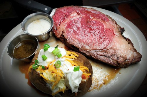 A plate featuring a juicy prime rib with sides of creamy sauce and a loaded baked potato topped with cheese and green onions.