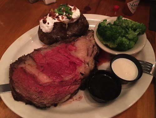A plate featuring a large slice of prime rib, a baked potato with toppings, broccoli, and dipping sauces.