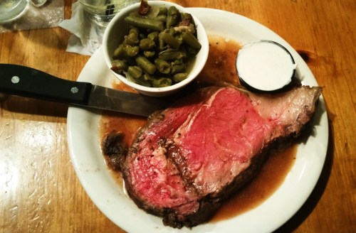 A plate with a large slice of prime rib, green beans, and a small cup of dipping sauce, set on a wooden table.