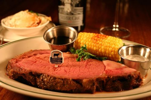 A plate of prime rib with corn, a baked potato, and dipping sauces, accompanied by a bottle of wine in the background.