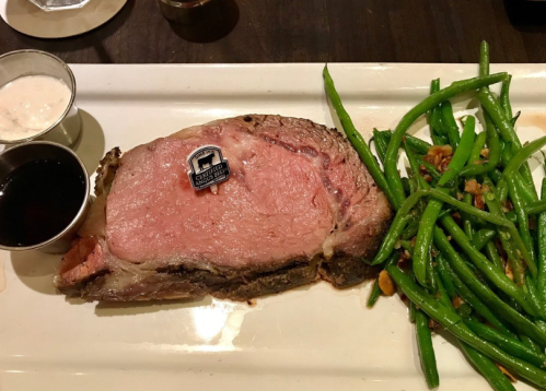 A plate featuring a slice of prime rib with a label, accompanied by green beans and two dipping sauces.