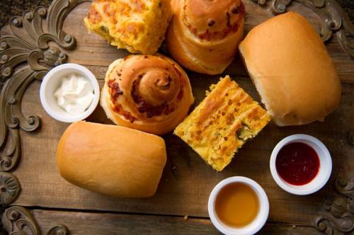 A wooden platter with various baked goods, including rolls, cornbread, and two small bowls of sauces.
