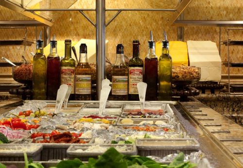 A buffet display featuring various toppings, oils, and vinegars, with fresh ingredients on ice and bottles in the background.