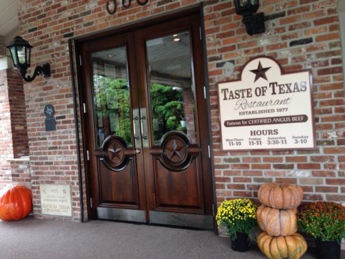 Entrance to Taste of Texas restaurant, featuring wooden doors, a sign with hours, and decorative pumpkins.