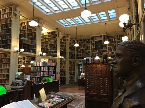 A spacious library with tall bookshelves, a bust statue, and green lamps, illuminated by natural light from skylights.