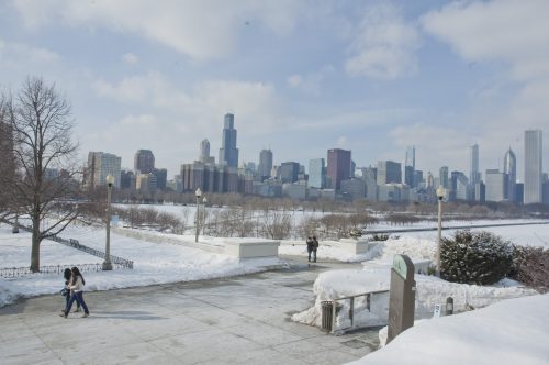 A snowy park with people walking, featuring a skyline of tall buildings in the background under a blue sky.