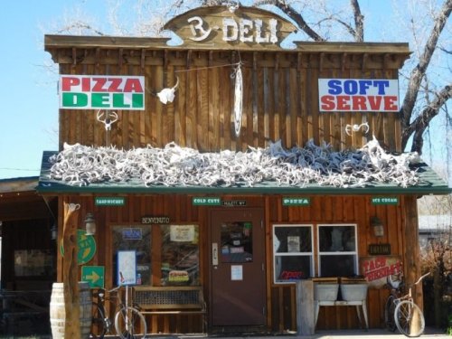 A rustic wooden deli with signs for pizza, soft serve, and sandwiches, featuring decorative antlers on the roof.