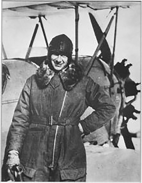 A smiling aviator in a fur-lined coat stands beside a vintage airplane, wearing a helmet and goggles.