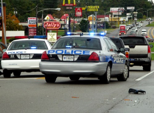 Two police cars with flashing lights on a busy road, with a few fast-food restaurants visible in the background.