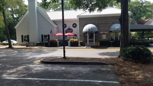 A restaurant with outdoor seating, umbrellas, and American flags, surrounded by trees and a paved parking area.