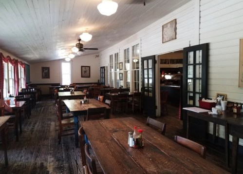 Cozy restaurant interior with wooden tables, chairs, and large windows, featuring rustic decor and warm lighting.