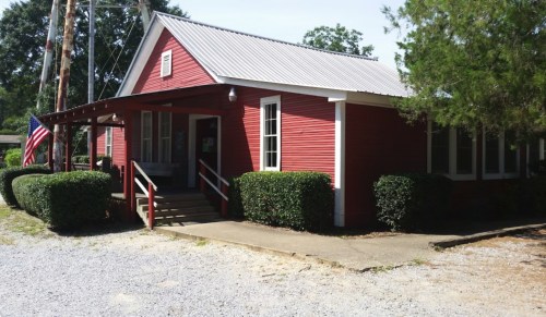 A small red building with a metal roof, surrounded by greenery and gravel, featuring a flagpole and steps at the entrance.