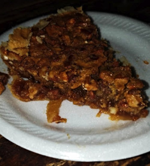 A slice of pecan pie on a white paper plate, featuring a golden-brown crust and a nutty filling.