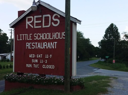 Sign for "Reds Little Schoolhouse Restaurant" with operating hours displayed, located near a rural road.