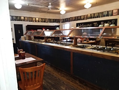 Interior of a rustic restaurant with a buffet counter, wooden tables, and framed photos on the walls.