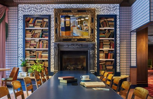 Cozy library setting with bookshelves, a large mirror, a fireplace, and a long table with books and a coffee cup.