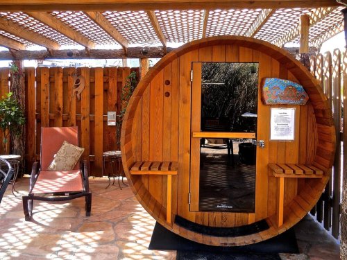 A wooden barrel sauna with a glass door, surrounded by a patio and lounge chair under a pergola.
