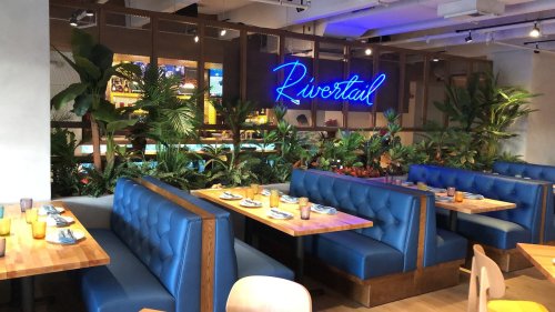 Interior of a restaurant featuring blue booths, wooden tables, and tropical plants, with a neon sign reading "Rivertail."