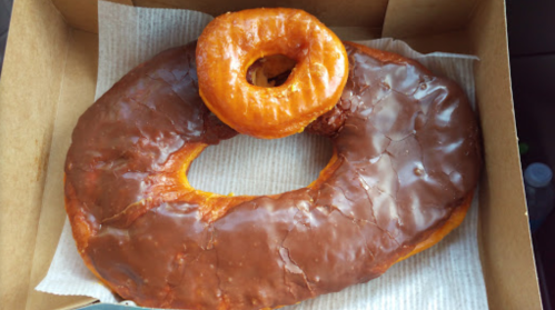 A large chocolate-glazed donut with a smaller glazed donut on top, both in a cardboard box lined with paper.