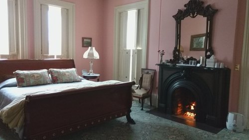 Cozy bedroom with a pink wall, wooden bed, vintage chair, and a fireplace with a mirror above it.