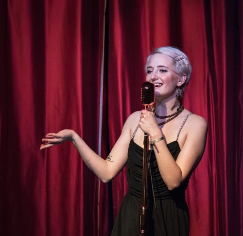 A singer in a black dress performs on stage with a vintage microphone, smiling against a backdrop of red curtains.