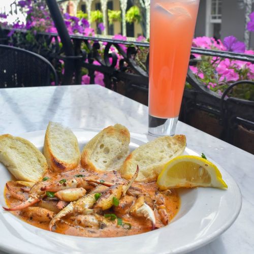 A plate of seafood in a creamy sauce, served with toasted bread and a lemon wedge, alongside a pink drink and flowers in the background.