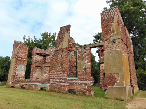 Ruins of a brick building with partially standing walls, surrounded by grass and trees under a cloudy sky.