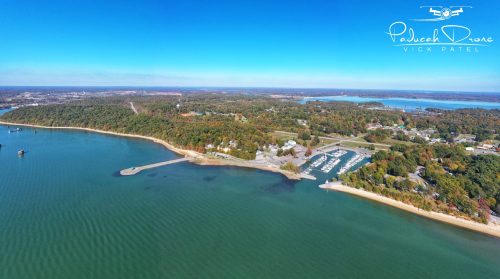 Aerial view of a serene lake with a marina, surrounded by colorful autumn foliage and clear blue skies.