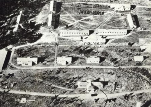 Aerial view of a large, abandoned facility with multiple buildings and open spaces, surrounded by trees.