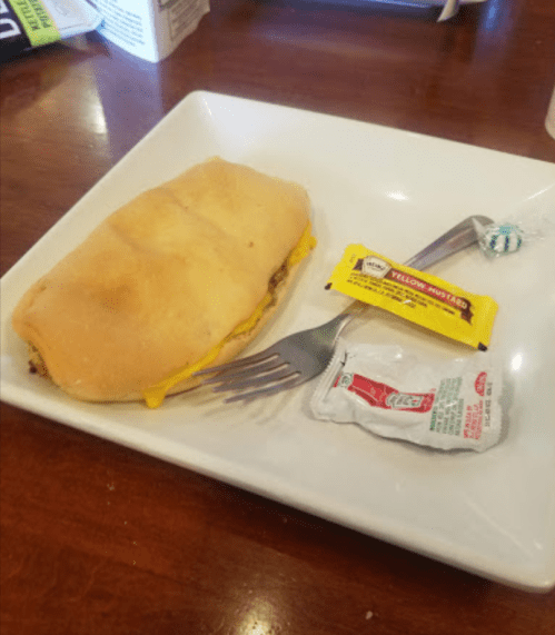 A sandwich with cheese and meat on a plate, accompanied by mustard and ketchup packets and a fork.