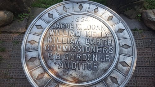 Metal plaque from 1894 listing names of commissioners and an auditor, set in a stone pathway.