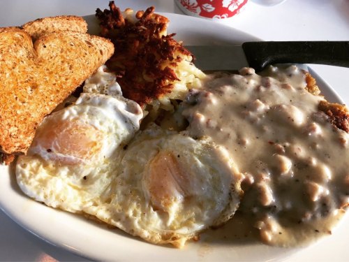A plate of breakfast featuring two fried eggs, hash browns, toast, and sausage gravy over biscuits.