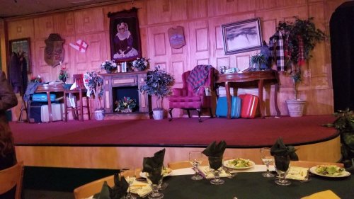 A stage set with two tables, a chair, and decorative plants, ready for a performance. Dining tables are in the foreground.