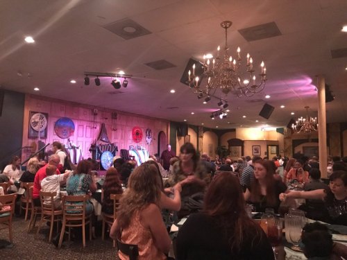 A lively restaurant scene with diners at tables, a stage in the background, and chandeliers overhead.