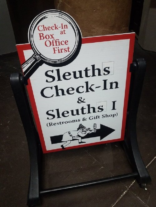 A sign directing visitors to check in at Sleuths, with arrows for restrooms and a gift shop.