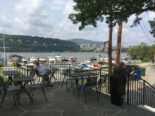 A serene riverside view with boats docked, a man sitting at a table, and lush greenery under a cloudy sky.
