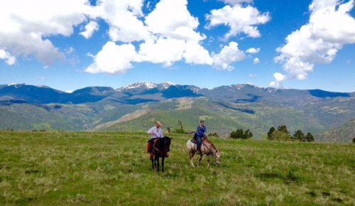 Two riders on horseback in a grassy field with mountains and clouds in the background.