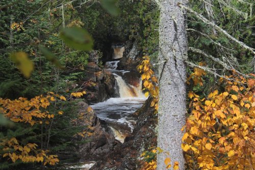 A serene waterfall cascades through rocky terrain, surrounded by vibrant autumn foliage and lush green trees.