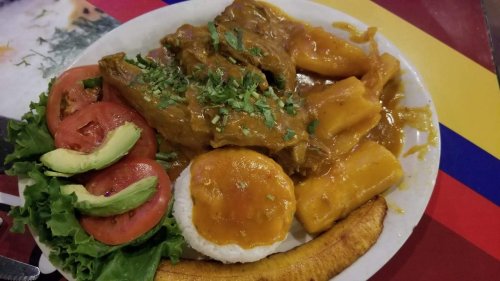A plate of traditional food featuring meat, plantains, yuca, rice, and fresh vegetables like tomatoes and avocado.