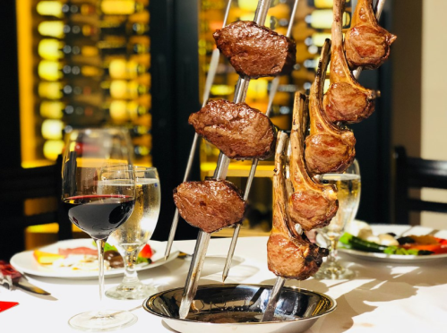 A platter of grilled meat skewers served with wine and water, set in an elegant dining environment.