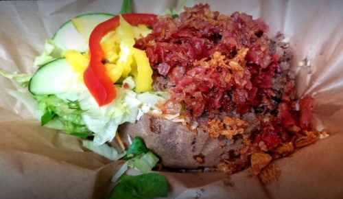 A loaded baked potato topped with bacon, lettuce, cucumber, and bell peppers, served in a paper bowl.