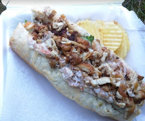 A large sandwich filled with shredded chicken, crispy toppings, and served with a side of potato chips.