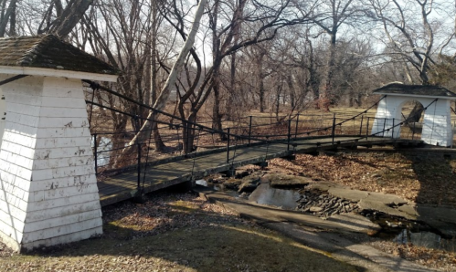 A wooden bridge connects two white structures over a stream, surrounded by bare trees and fallen leaves.