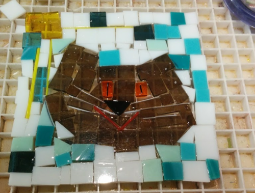 A colorful mosaic of a cat's face, featuring brown, white, and turquoise tiles with orange eyes and a red mouth.
